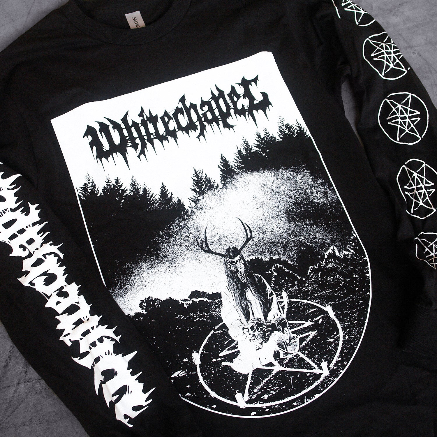 close up image of a black long sleeve tee shirt laid on a concrete floor. front of shirt has full body print in white that says whitechapel at the top over a scene of a ritual in the woods with a pentagram. left sleeve says whitechapel and right sleeve has five pentagrams