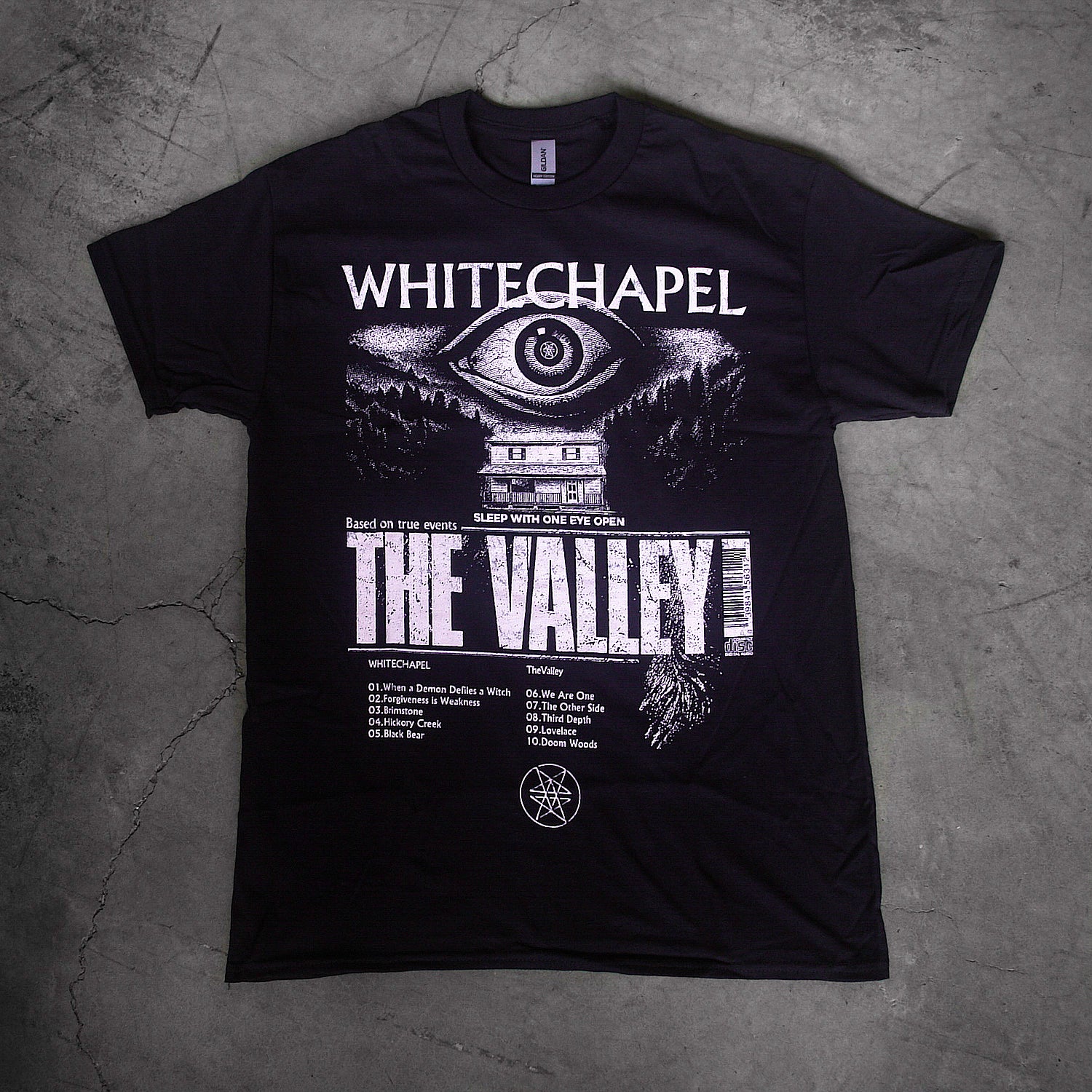 image of a black tee shirt laid flat on a concrete floor. tee has full body print in white of a giant eye above a house. at the top says whitechapel and below says the valley