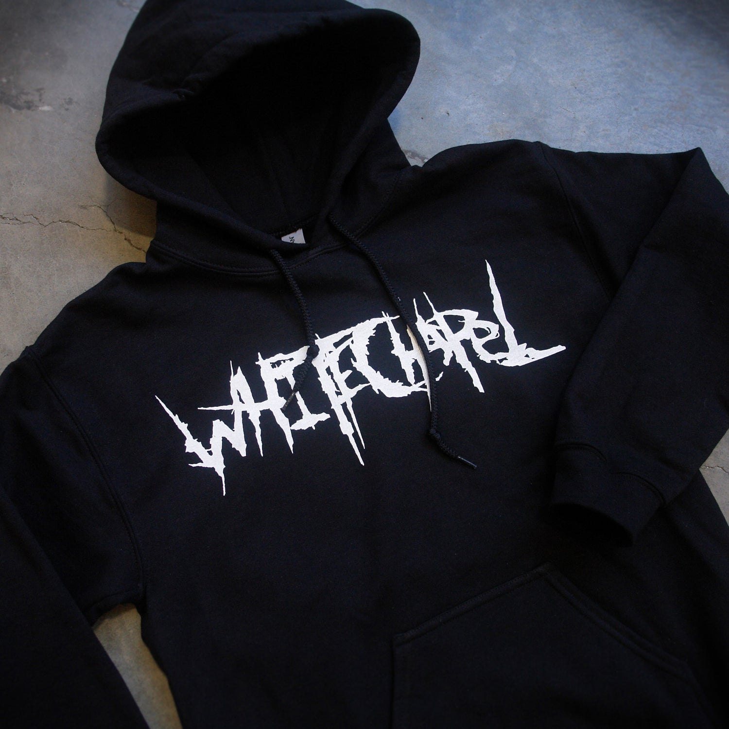 image of a black pullover hoodie laid flat on a concrete floor. hoodie has center chest print in white that says whitechapel