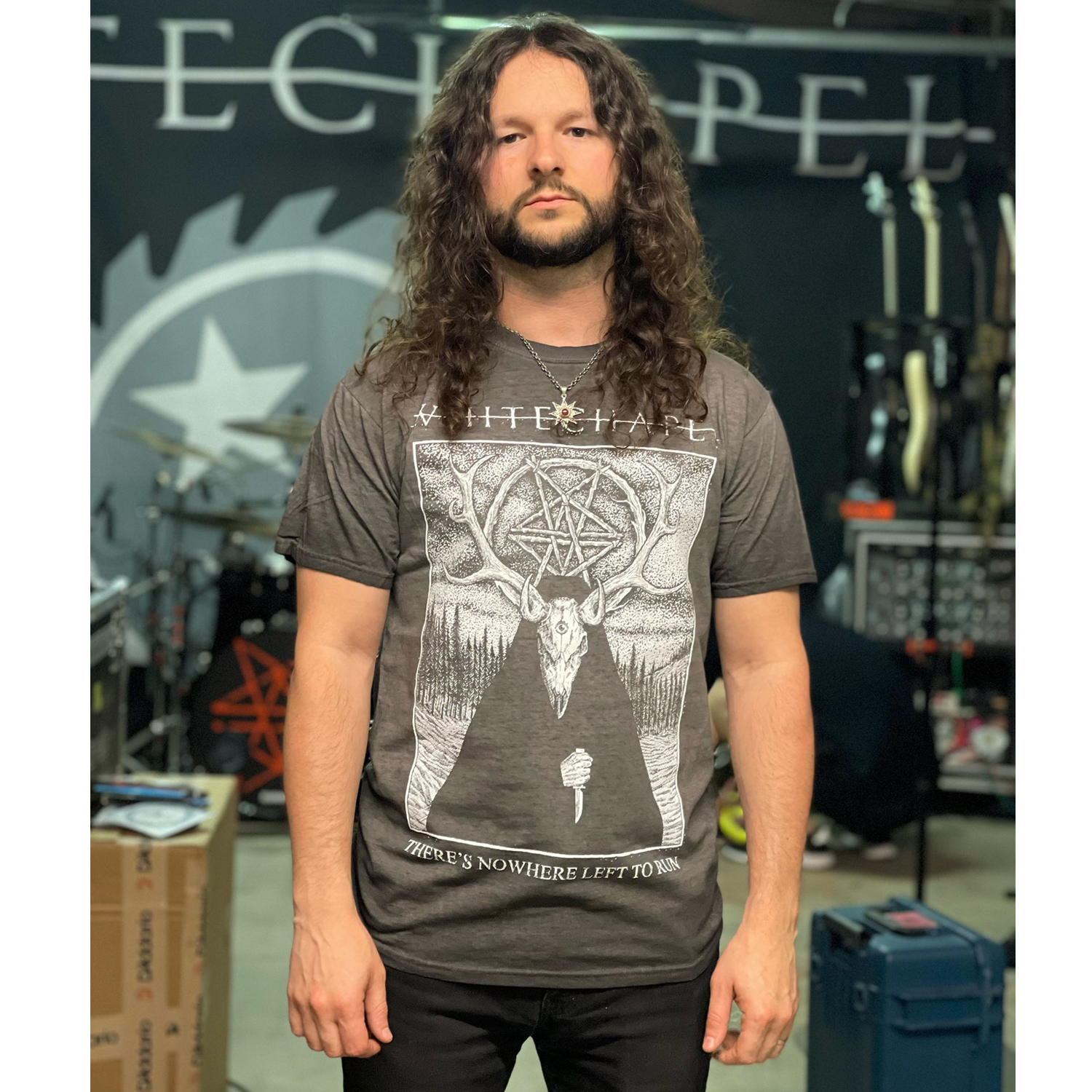 image of a man with curly long hair wearing an oil washed black t standing in the whitechapel music studio. tee has full body print in white of a deer wearing a cloak outside in the woods. at the top says whitechapel and the bottom says there's nowhere left to run