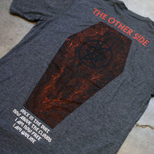 Load image into Gallery viewer, up close Image of the back of a heather charcoal tshirt against a grey concrete background. the top of the tee near the shoulders reads &quot;the other side&quot;. it is in red text. below this is a graphic of a coffin with a black double pentagram (two stars mirroring and slightly overlapping each other) on the center of it. Below this in white text reads once in the dirt, now above the clouds. i am now free, i am god like.
