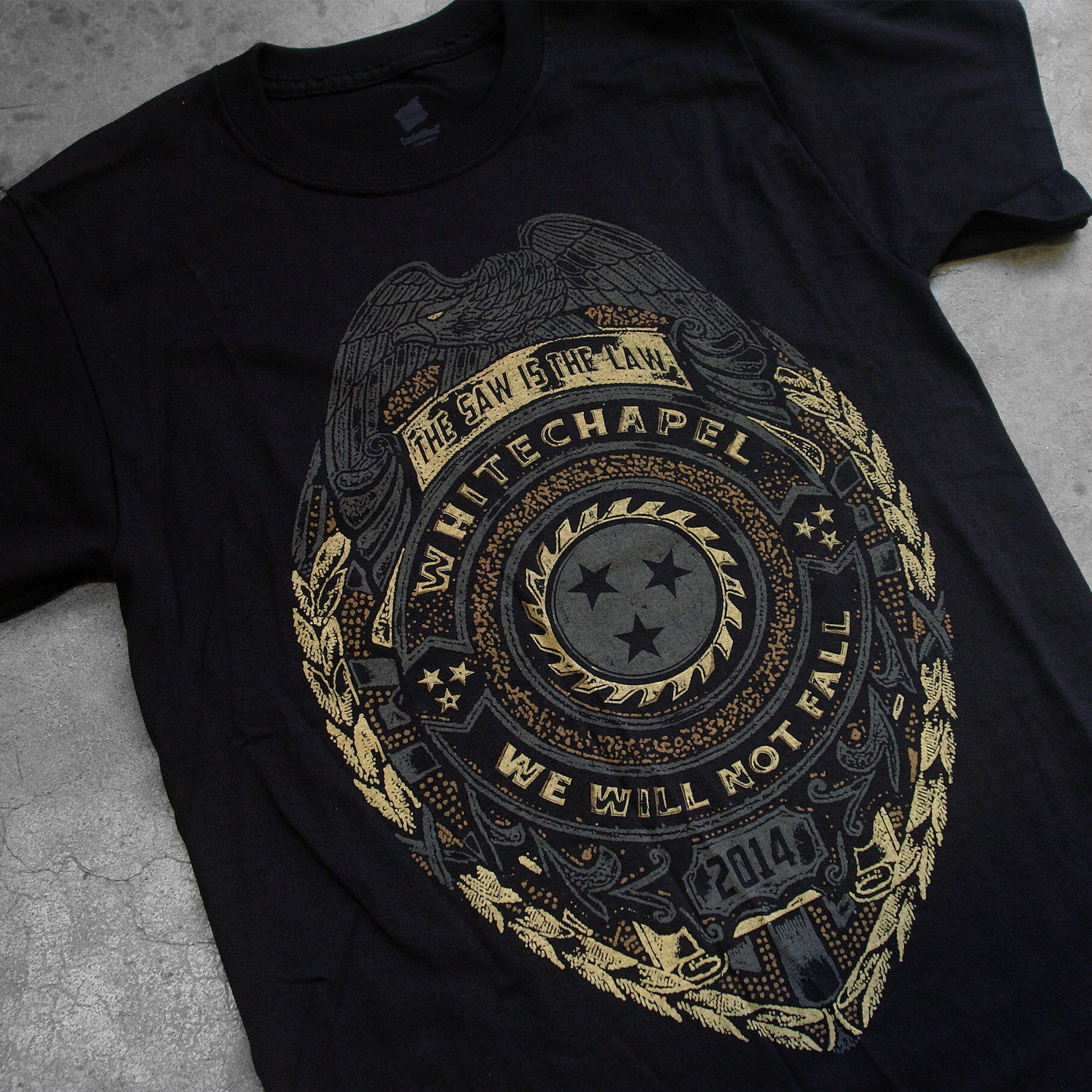 close up, angled Image of a black tshirt against a concrete background. The shirt is a graphic of a badge. There is a bird at the top, and below that it says the saw is the law. the center of the badge has a circle with a sawblade with three stars on the inside of the saw blade. Wrapped around the circle reads "whitechapel, we will not fall". Below this it says 2014. The graphic is a faded grey/gold and black color.