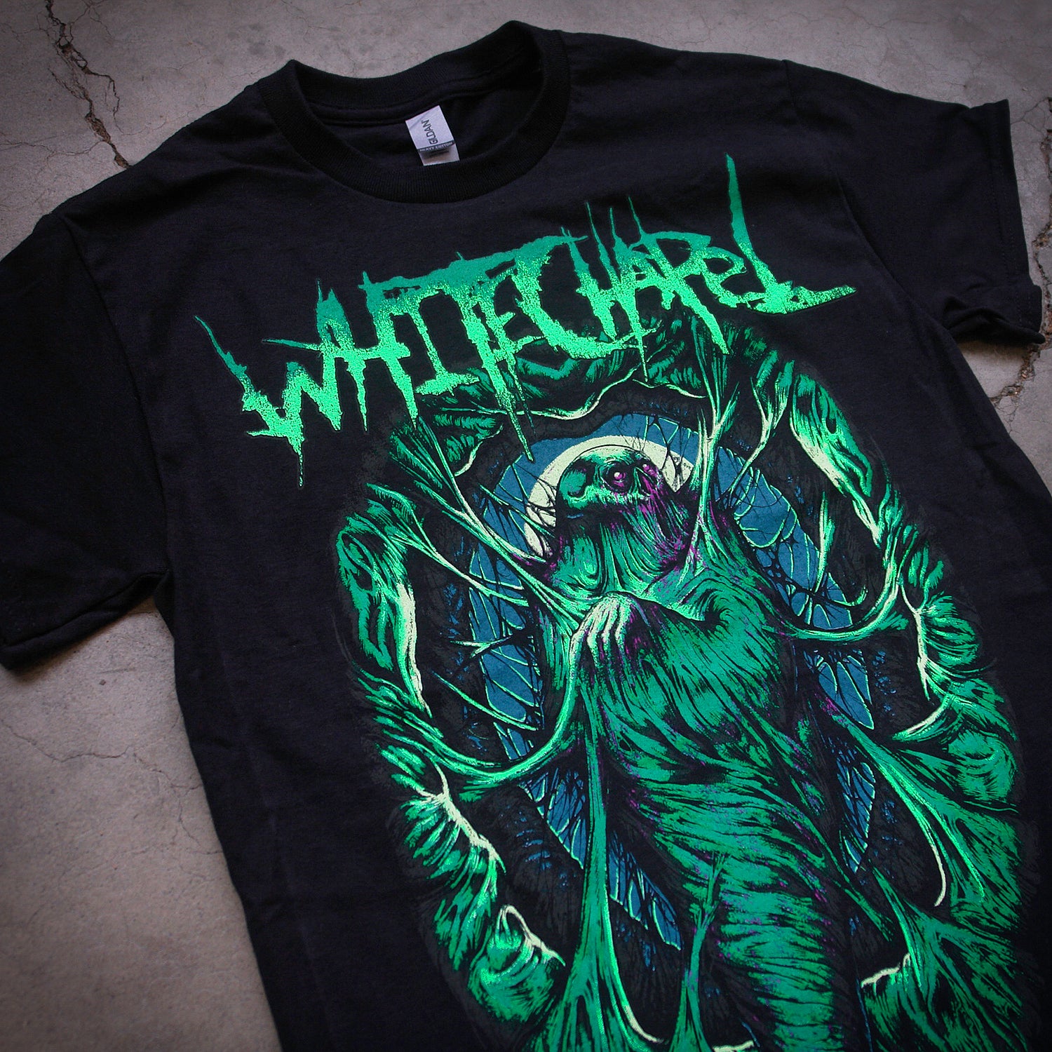 close up, angled image of a black tee shirt laid flat on a cracked concrete floor. tee has full body print of a green cocoon with a skeleton inside. at the top says whitechapel