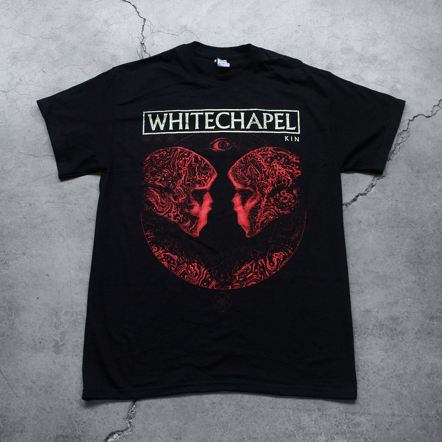 Image of the front of a black tshirt on a grey concrete background. The front shows two skulls/skeletal figures from the shoulders up looking at one another- made up of red and black oil. This is outlined in a thin semi circle. Above the heads of the skulls is a red opened eye. Above this in white text with a white rectangular outline is the word 