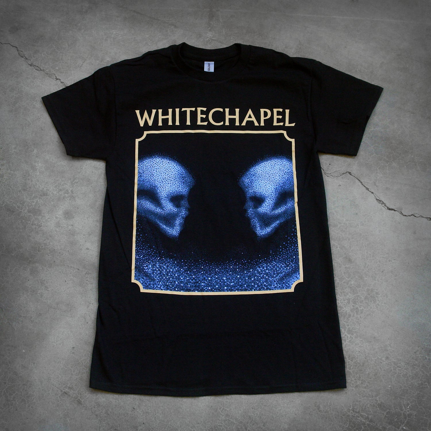 image of the front of a black tee shirt on a cracked concrete background. The front shows two skulls looking at one another- made up of blue and black static. The static is beneath them too. This is outlined in a tan square. Above the square in tan text reads 