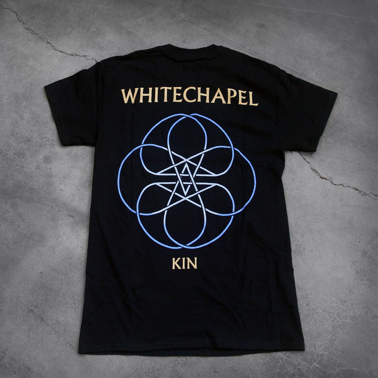 image of the back of a black tee shirt on a cracked concrete background. The back reads "whitechapel" across the shoulders in tan text. Below this is an abstract thin lined pattern in white and blue. It looks like a circle with lines and stars in the center. Below this in tan text reads "kin".