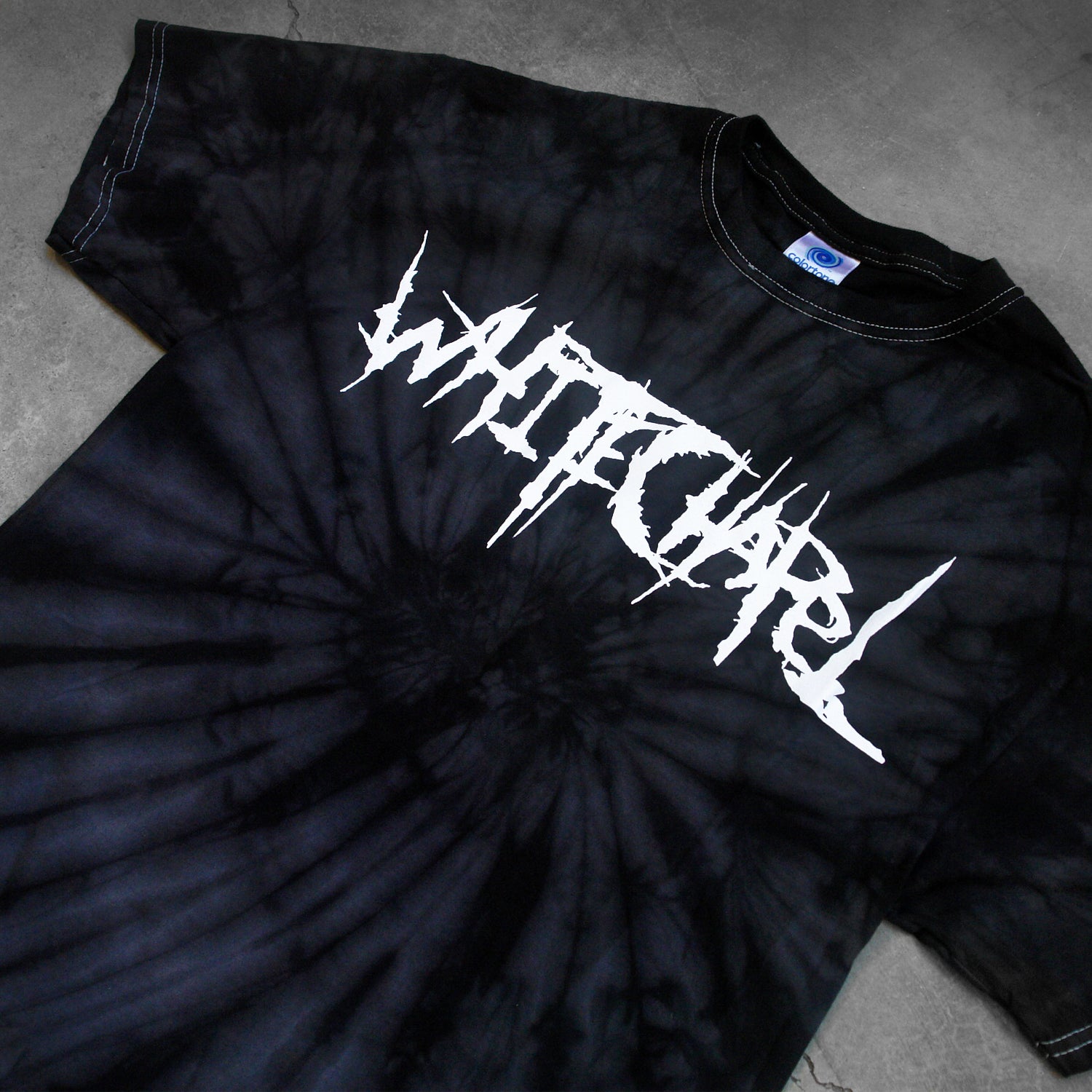 close up, angled image of a black spider tie dye tee shirt on a cracked concrete background. tee has front center chest print across in white that says Whitechapel