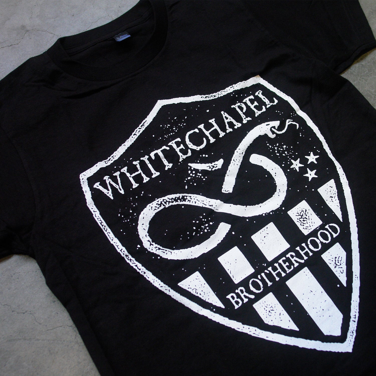 Image of a black tshirt against a grey concrete background. There is a graphic of a white badge on the shirt, inside this badge in white text reads "whitechapel". Below this is a graphic of a snake with its tongue out. There are three small stars next to the snake. Below this are white stripes, and in between the stripes in the middle reads "brotherhood".
