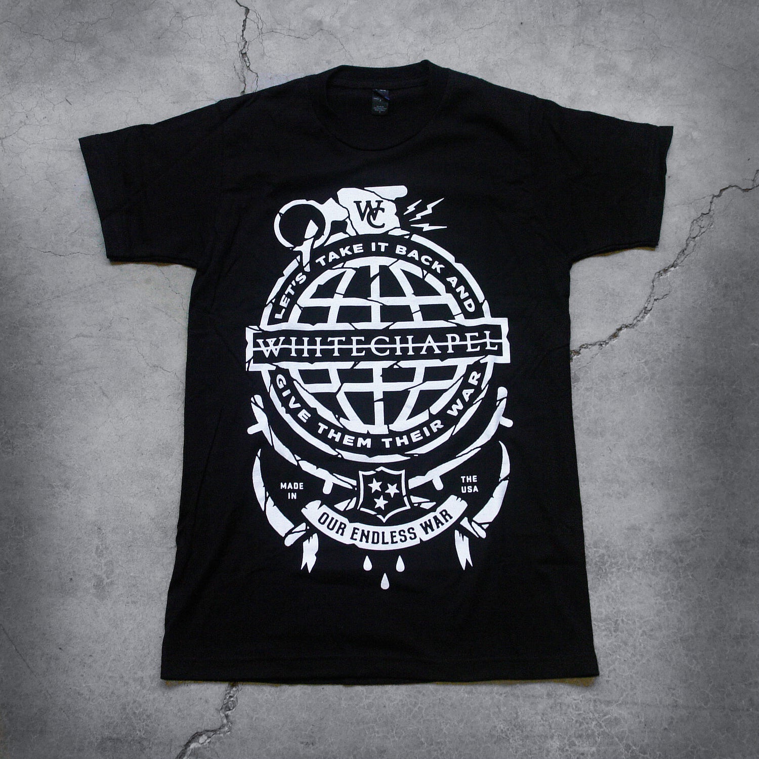 Image of the front of a black tshirt against a grey concrete background. There is a graphic of a white grenade and in there is a globe in the center of it with the text 