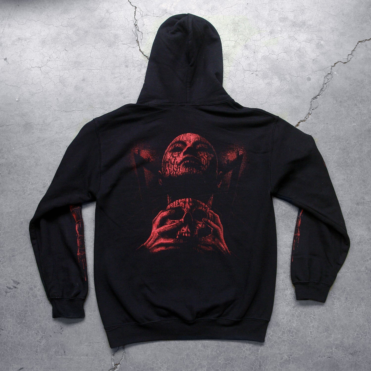  Image of the back of a black hoodie against a concrete grey background.  There is a red graphic of a person's face with their eyes closed, head up. They have blood on them and are clutching a skull.  The sleeves feature red graphics that are abstract and drip blood.