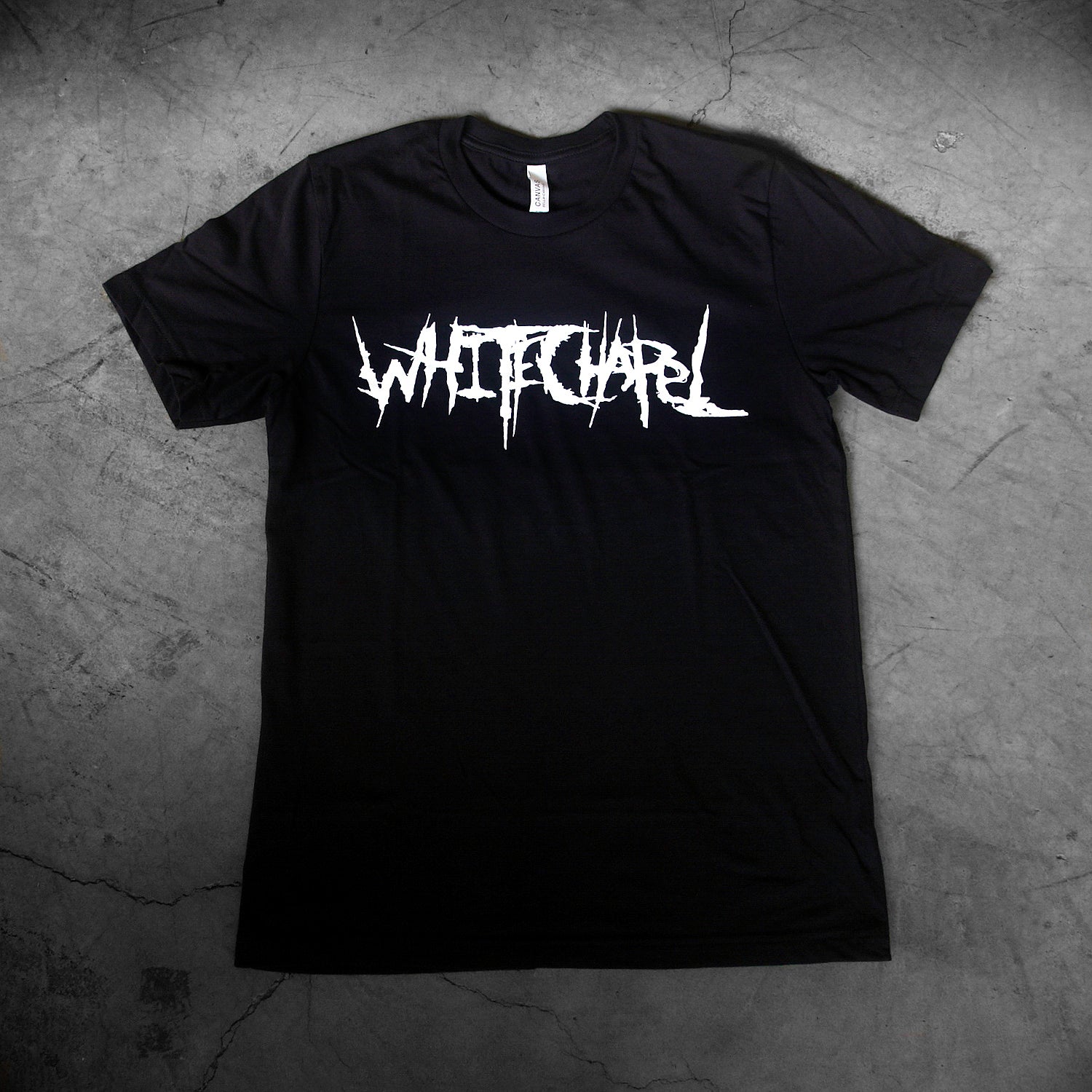 image of a black tee shirt laid flat on a concrete floor. tee has center chest print in white that says whitechapel