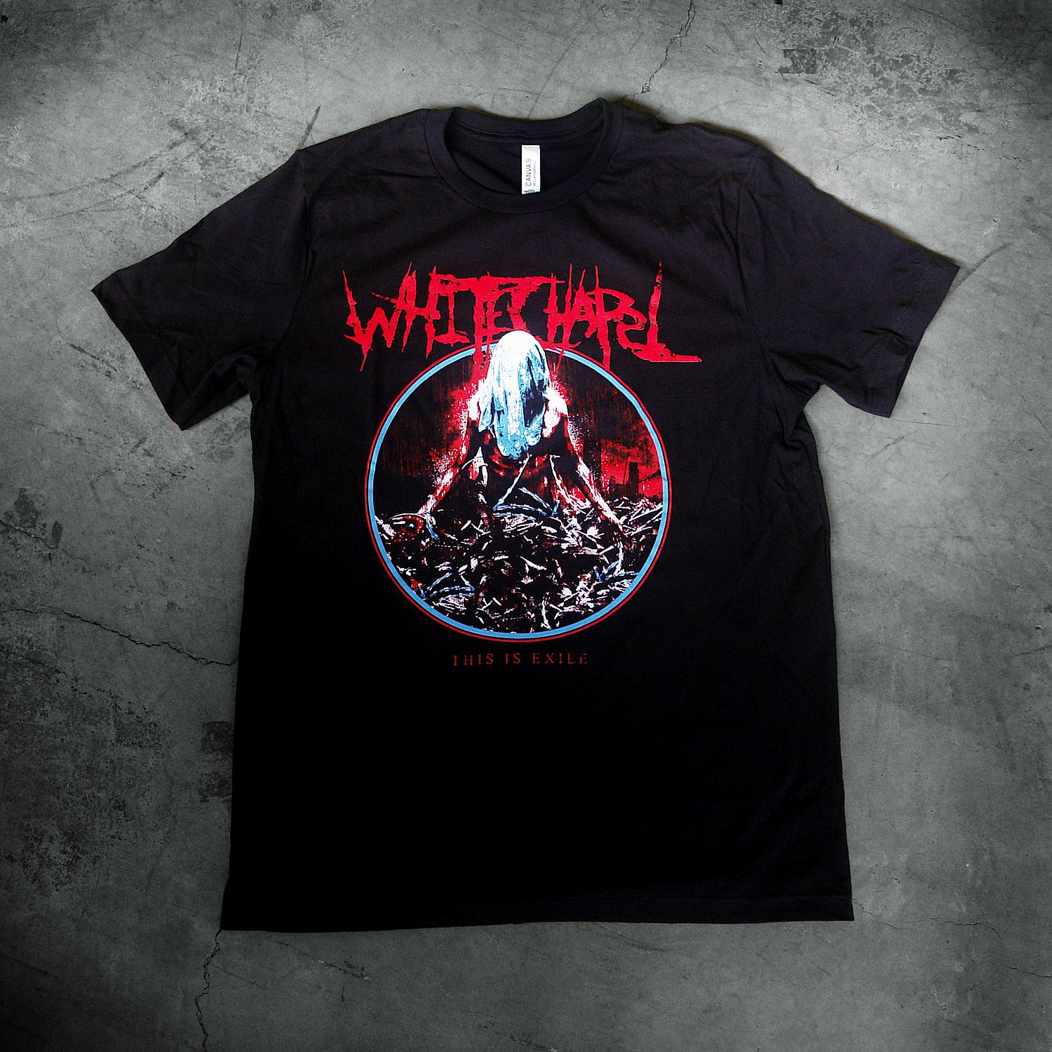 image of a black tee shirt laid flat on a concrete floor. tee has center chest print. at the top in red says whitechapel. below is a circle, with an image of a man with a sheet over his head wrapped in barbed wire. at the bottom says this is exile.