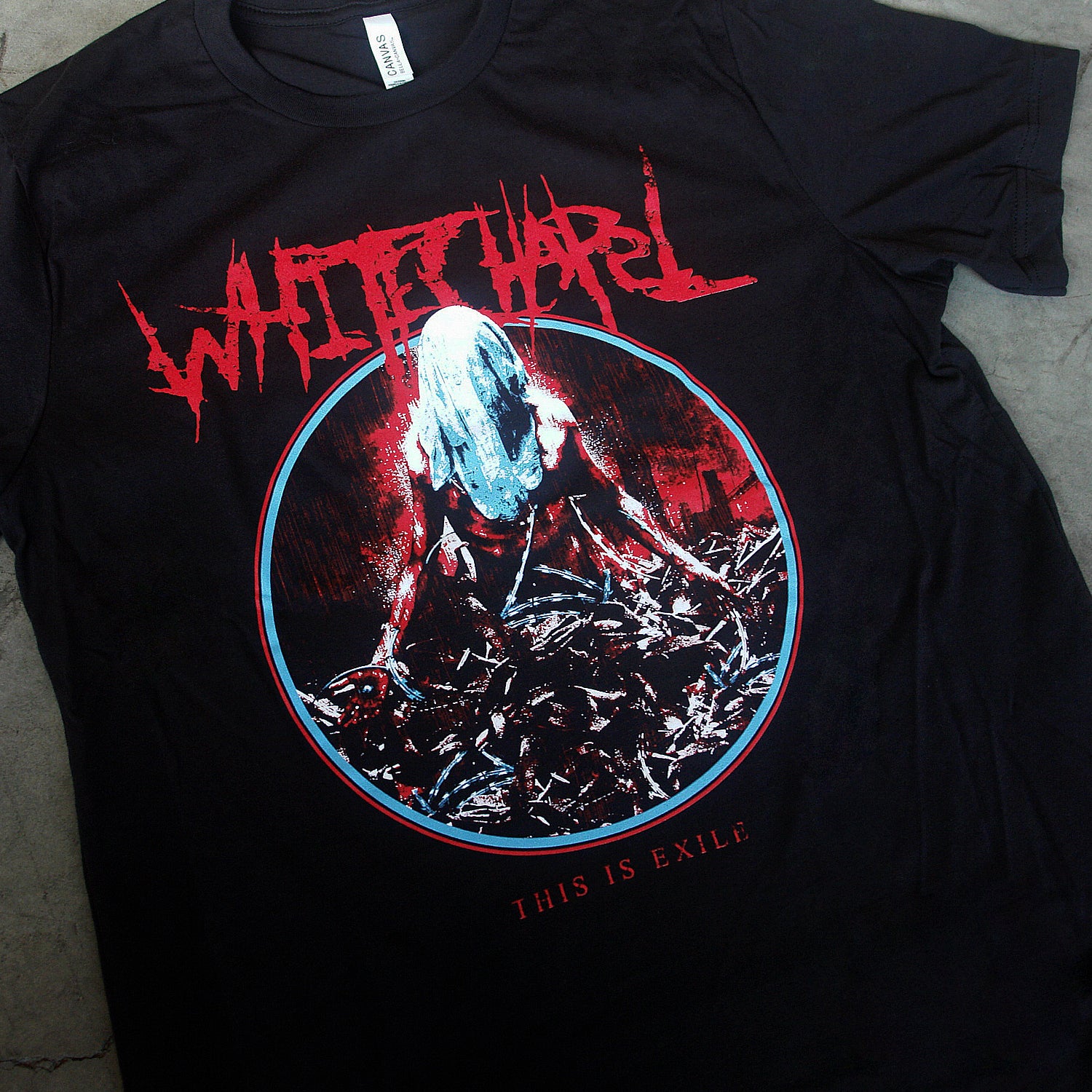 close up, angled image of a black tee shirt laid flat on a concrete floor. tee has center chest print. at the top in red says whitechapel. below is a circle, with an image of a man with a sheet over his head wrapped in barbed wire. at the bottom says this is exile.