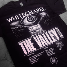 Load image into Gallery viewer, close up, angled image of a black tee shirt laid flat on a concrete floor. tee has full body print in white of a giant eye above a house. at the top says whitechapel and below says the valley
