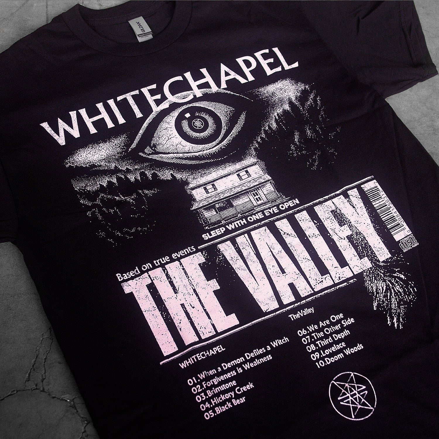 close up, angled image of a black tee shirt laid flat on a concrete floor. tee has full body print in white of a giant eye above a house. at the top says whitechapel and below says the valley