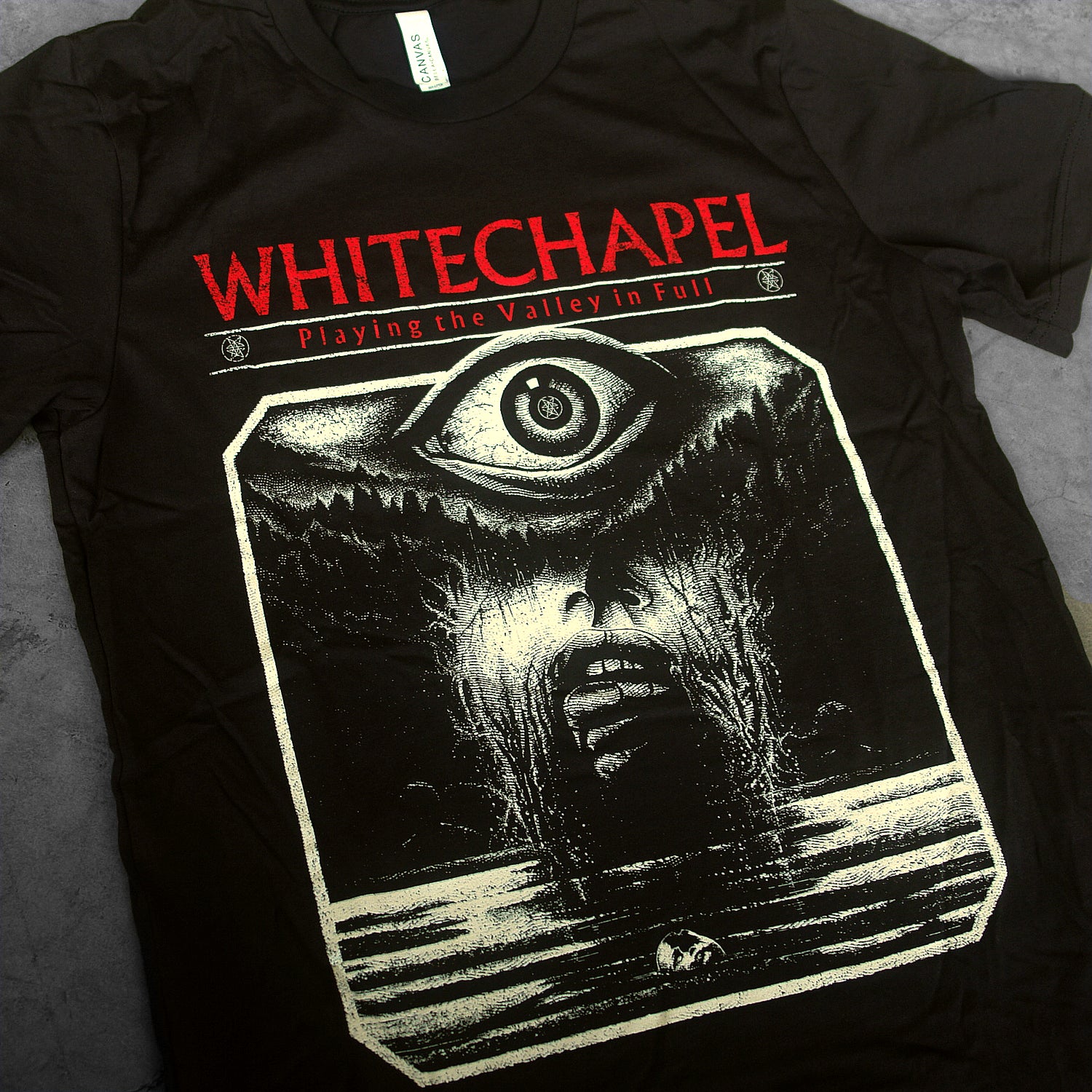 close up, angled image of a black tee shirt laid flat on a concrete floor. tee has full print of an eye coming of out water. at the top in red says whitechapel playing the valley in full