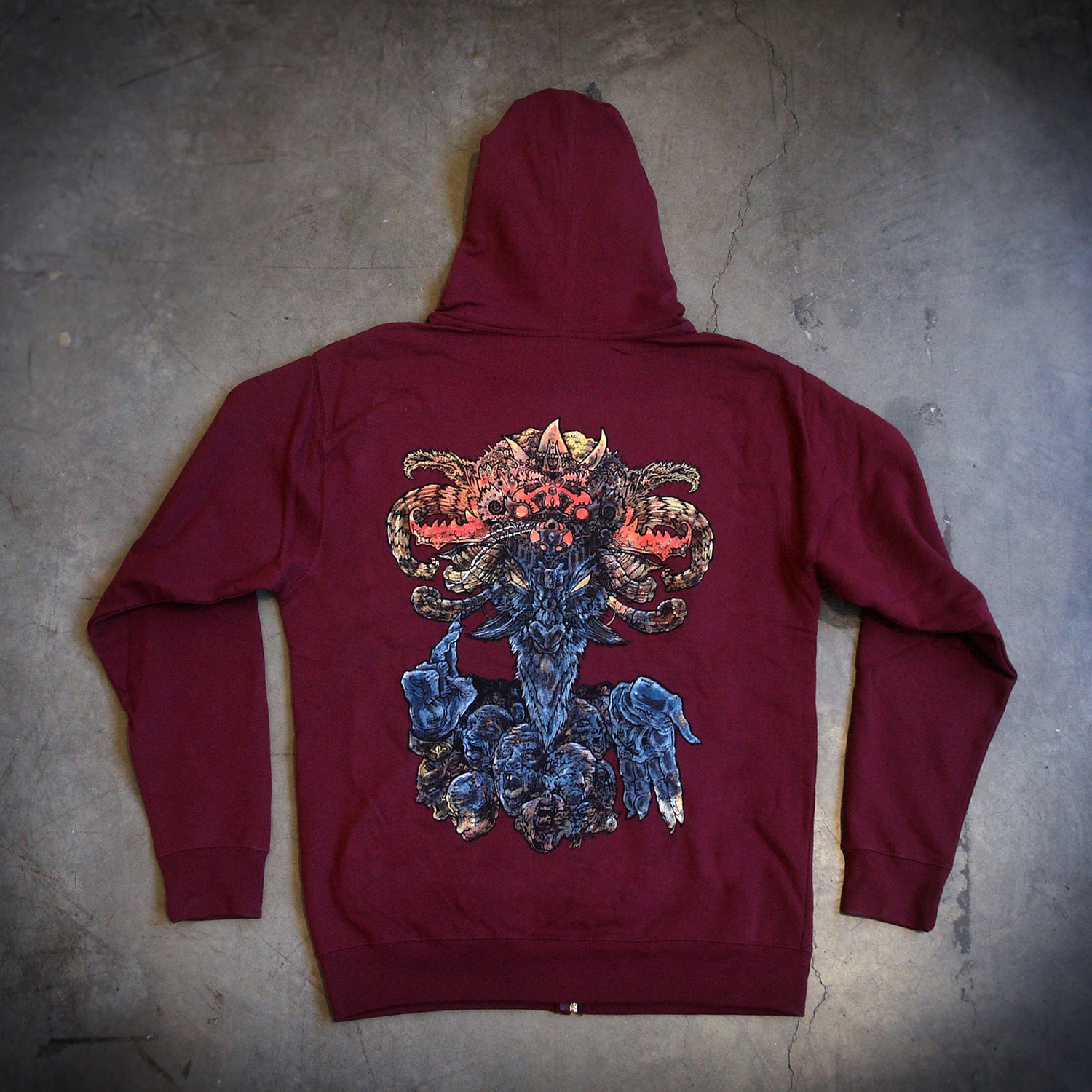 image of the back of a maroon hoodie laid flat on a concrete floor. the hoodie has a full back print of a goat like god figure