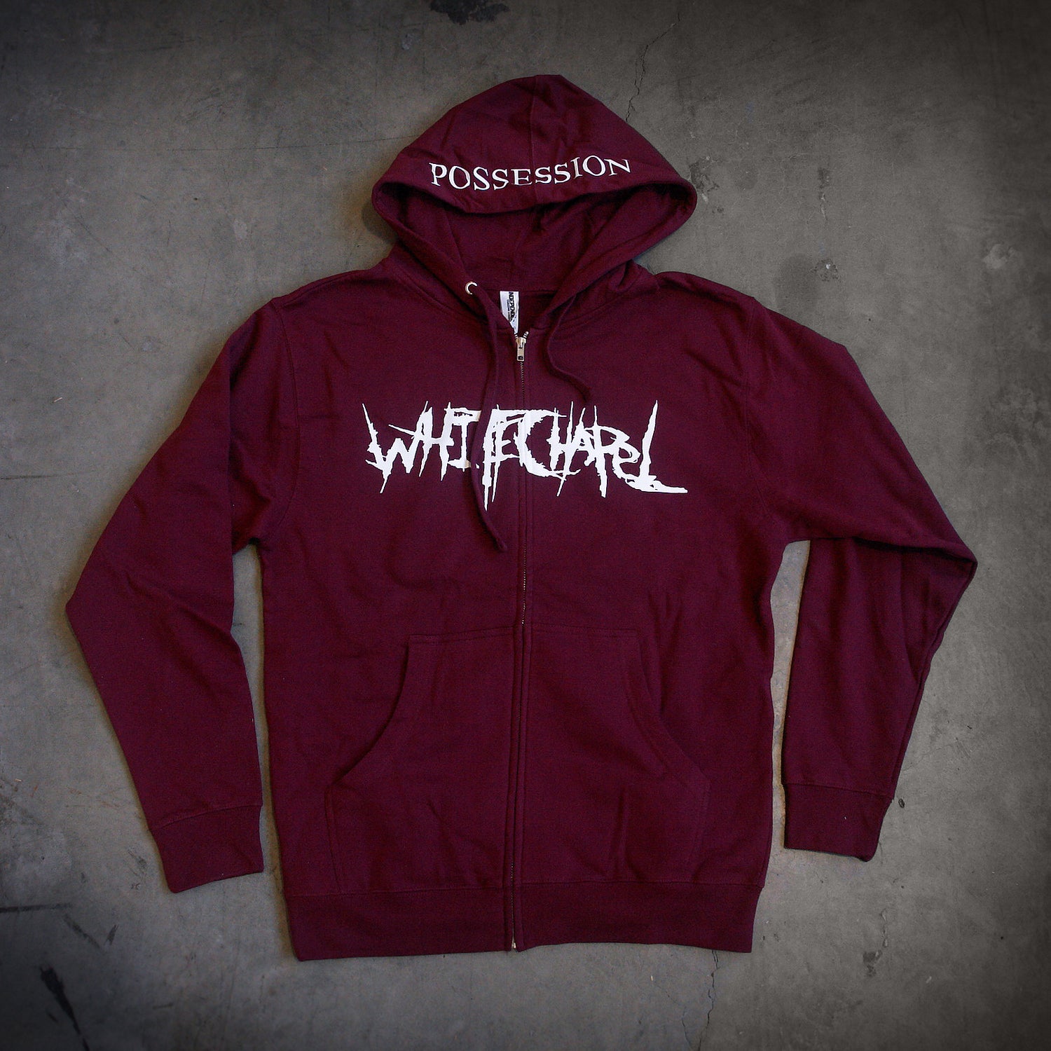 image of a maroon zip up hoodie laid flat on a concrete floor.  front of the hoodie is on the left and has a white print across the chest that says whitechapel. the hood is in the middle and shows a white print across the brim that says possession.