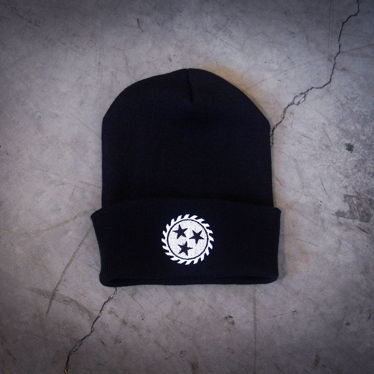 image of a black winter beanie laid pn a concrete floor. beanie has white embroidery on the front center cuff of a sawblade with three stars