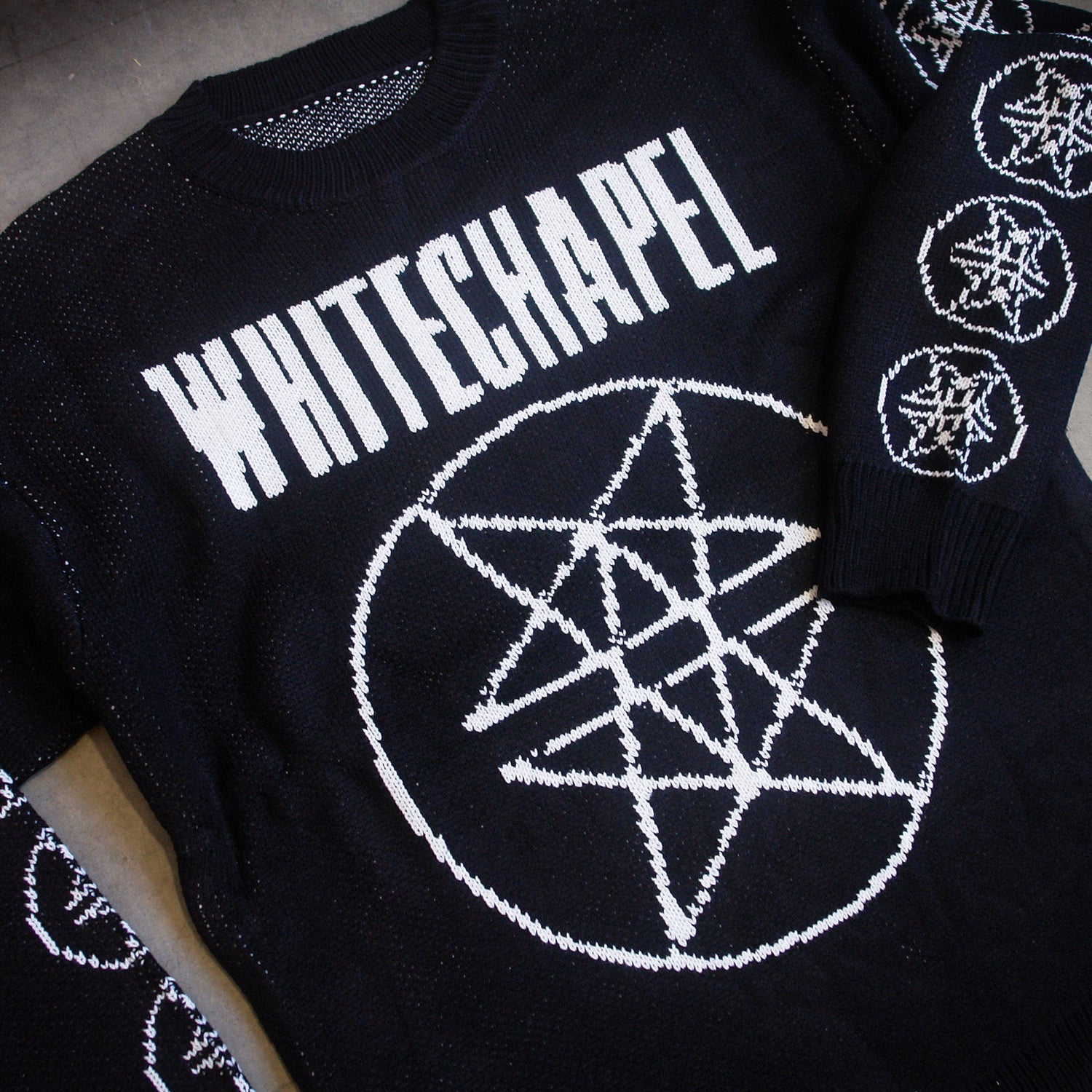 image of a custom black woven sweater laid flat on a concrete floor. sweater has white thread. the front has a full body double pentagram in the center and says whitechapel at the top across the chest. each sleeve has six double pentagrams 