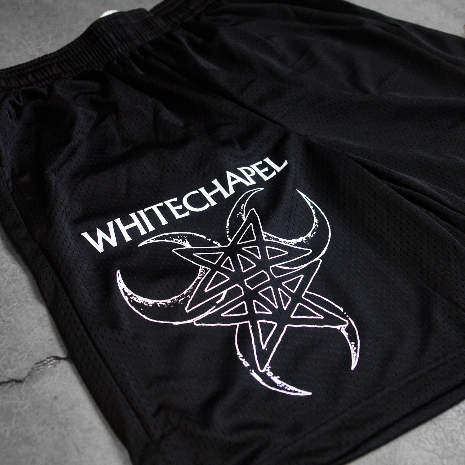 close up Image of black athletic shorts against a grey concrete background. The right bottom of the leg of the shorts says "whitechapel" across the leg in white text. Below that is a graphic of three cresent moons outlined in white- one facing the left, one the right, and one straight down. In the center of that is a double pentagram- two stars mirroring and slightly overlapping each other, also in white. 