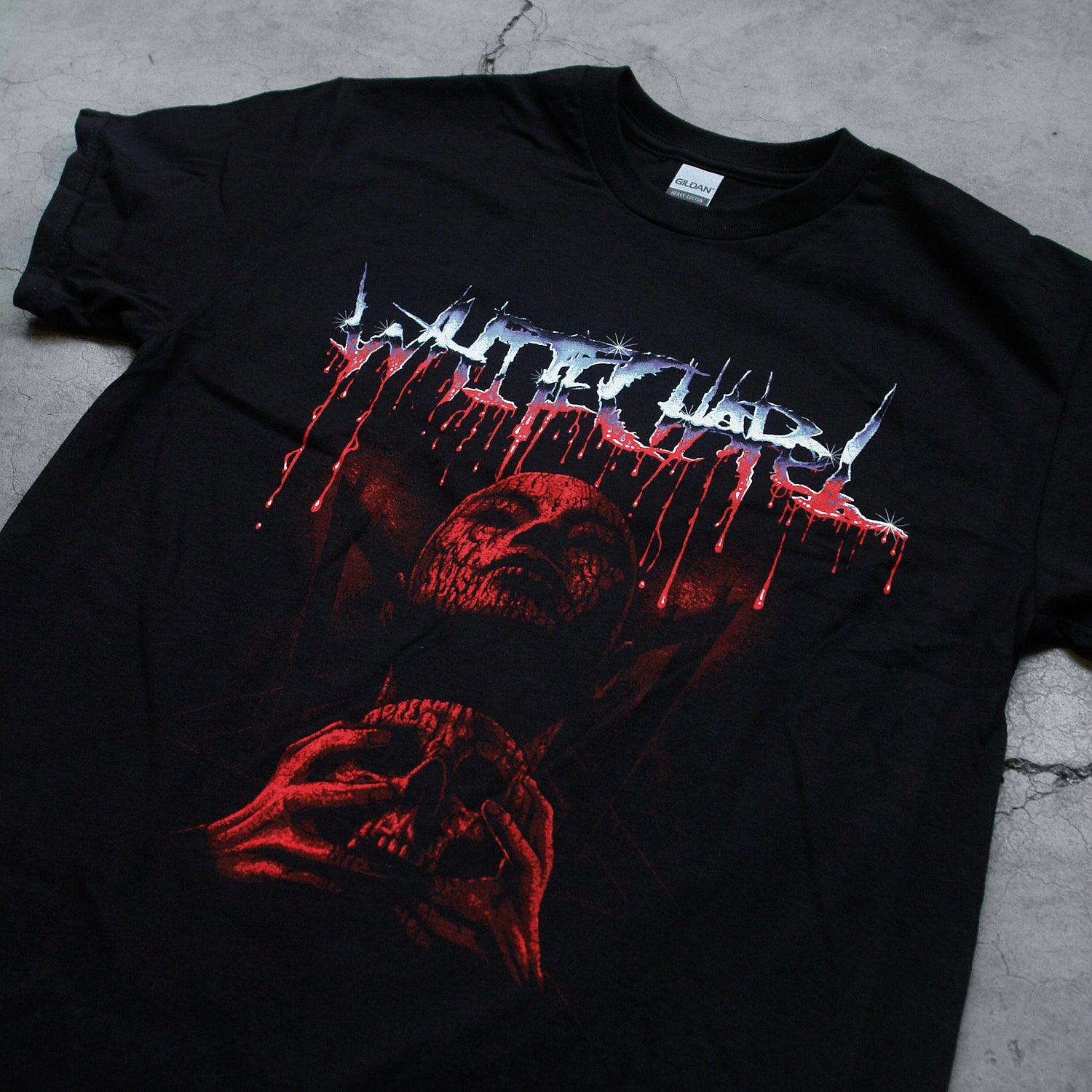 close up Image of the front of a black tshirt against a concrete grey background. Across the chest in a blue to white to red gradient color reads "whitechapel". This is in a heavy metal font and the red is dripping. Below this is a red graphic of a person's face with their eyes closed, head up. They have blood on them and are clutching a skull.
