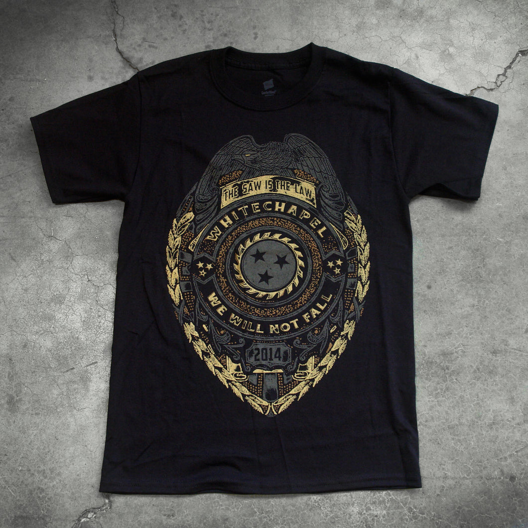 Image of a black tshirt against a concrete background. The shirt is a graphic of a badge. There is a bird at the top, and below that it says the saw is the law. the center of the badge has a circle with a sawblade with three stars on the inside of the saw blade. Wrapped around the circle reads 