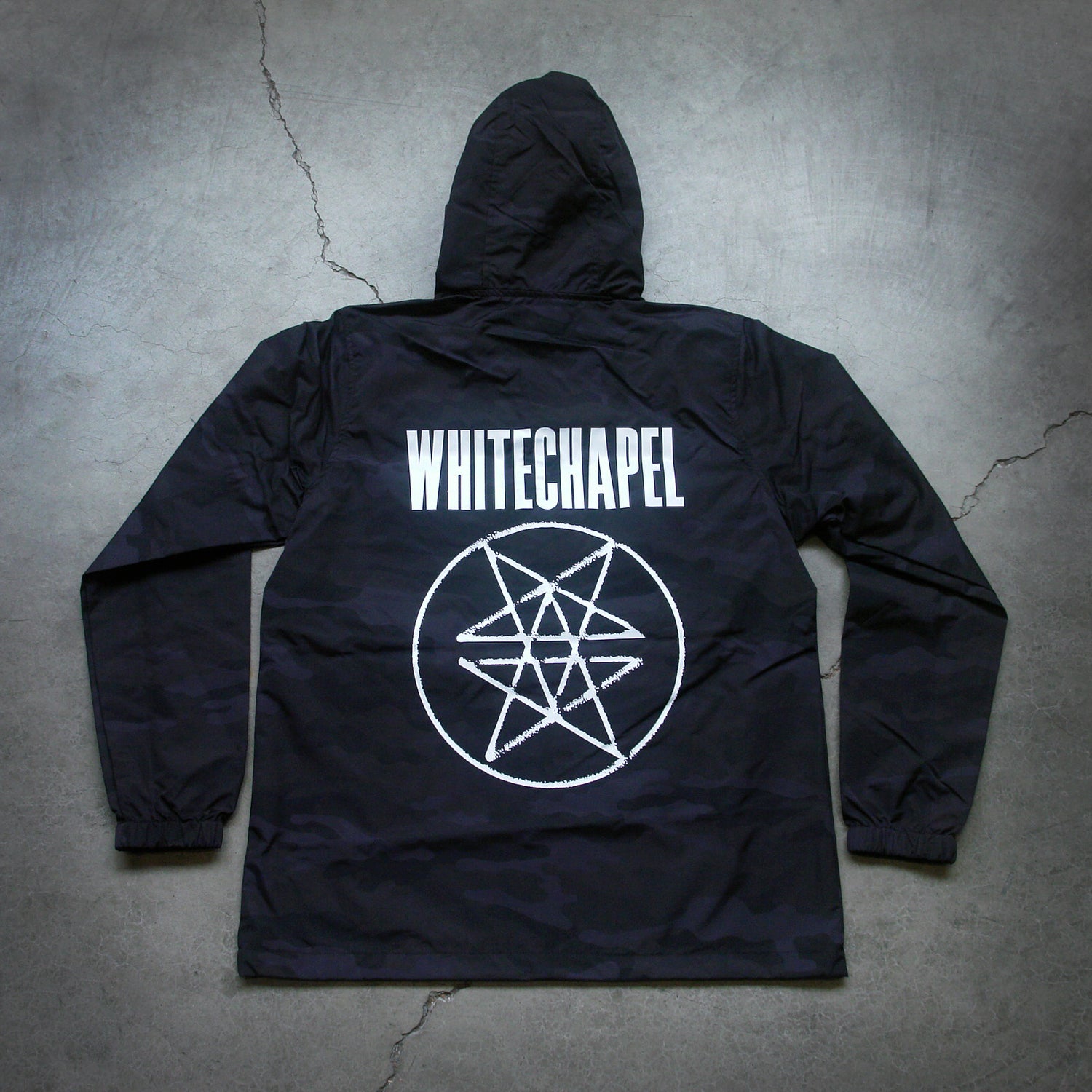 Image of the back of a black pullover windbreaker against a grey concrete background. Across the shoulder area of the jacket in big white letters says "whitechapel". Below that is what is called a double pentagram, it is a white circle with two stars mirroring each other on the inside of the circle. 