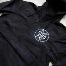 Load image into Gallery viewer, close up Image of the front of a black pullover windbreaker against a grey concrete background. The left chest has what is called a double pentagram, it is a white circle with two stars mirroring each other on the inside of the circle. There is a quarter zip on the windbreaker and a large kangaroo pouch in the center. 
