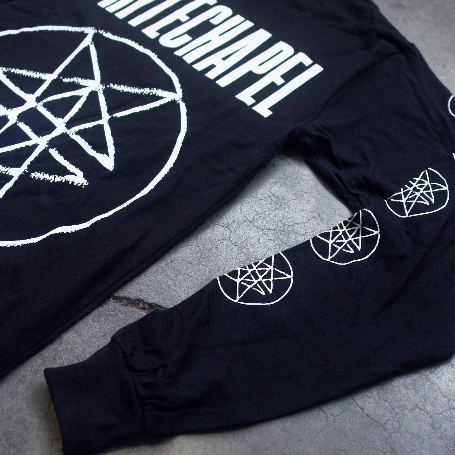 Close up Image of the front of a black longsleeve against a grey background. Across the chest in big white letters says "whitechapel". Below that is what is what is called a large double pentagram-  it is a white circle with two stars mirroring each other on the inside of the circle. Both sleeves also have double pentagrams going down the sleeves from the top to the bottom.