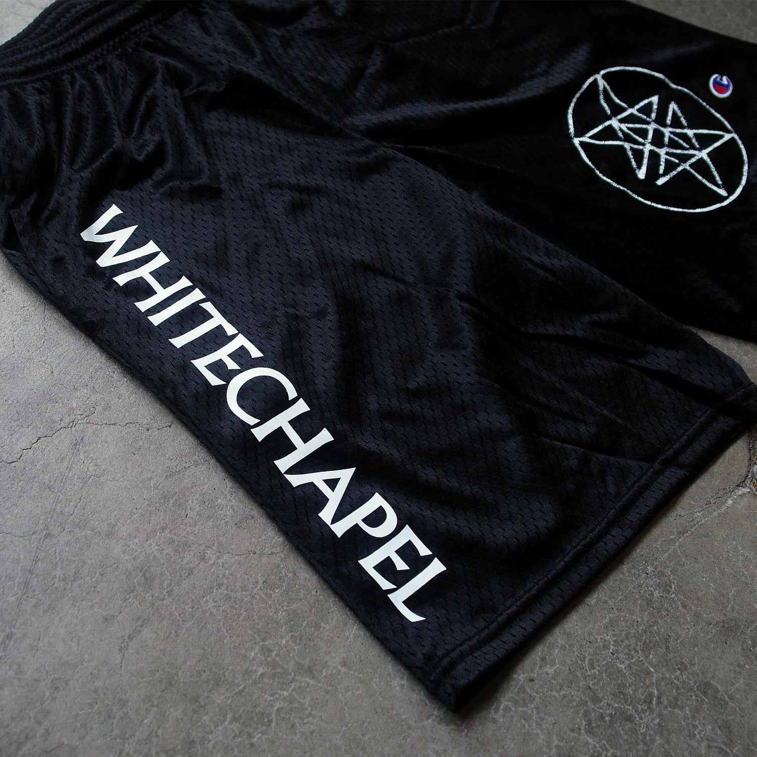 close up Image of black shorts against a grey concrete background. The shorts are athletic style with a drawstring. Descending down the right leg in white text says "whitechapel". The bottom of the left leg features a double pentagram- a white circle with two stars that are mirroring and overlapping each other in the center of it. The champion "C" red white and blue logo is next to this. 