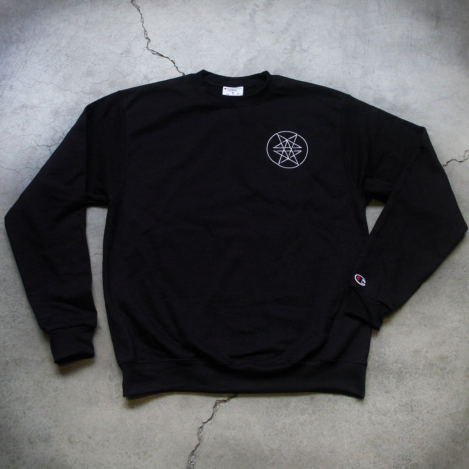 Image of the front of a black crewneck against a grey concrete background. The left chest has what is called a double pentagram, it is a white circle with two stars mirroring and slightly overlapping each other on the inside of the circle. The left sleeve has the champion 