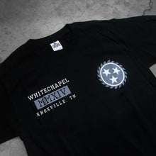 Load image into Gallery viewer, close up, angled image of the front of a black tee shirt on a concrete background. the front of the tshirt has a grey sawblade with three white stars in the center of the sawblade on the left chest. The right chest says whitechapel, mmxiv, knoxville tn in a grey white text.
