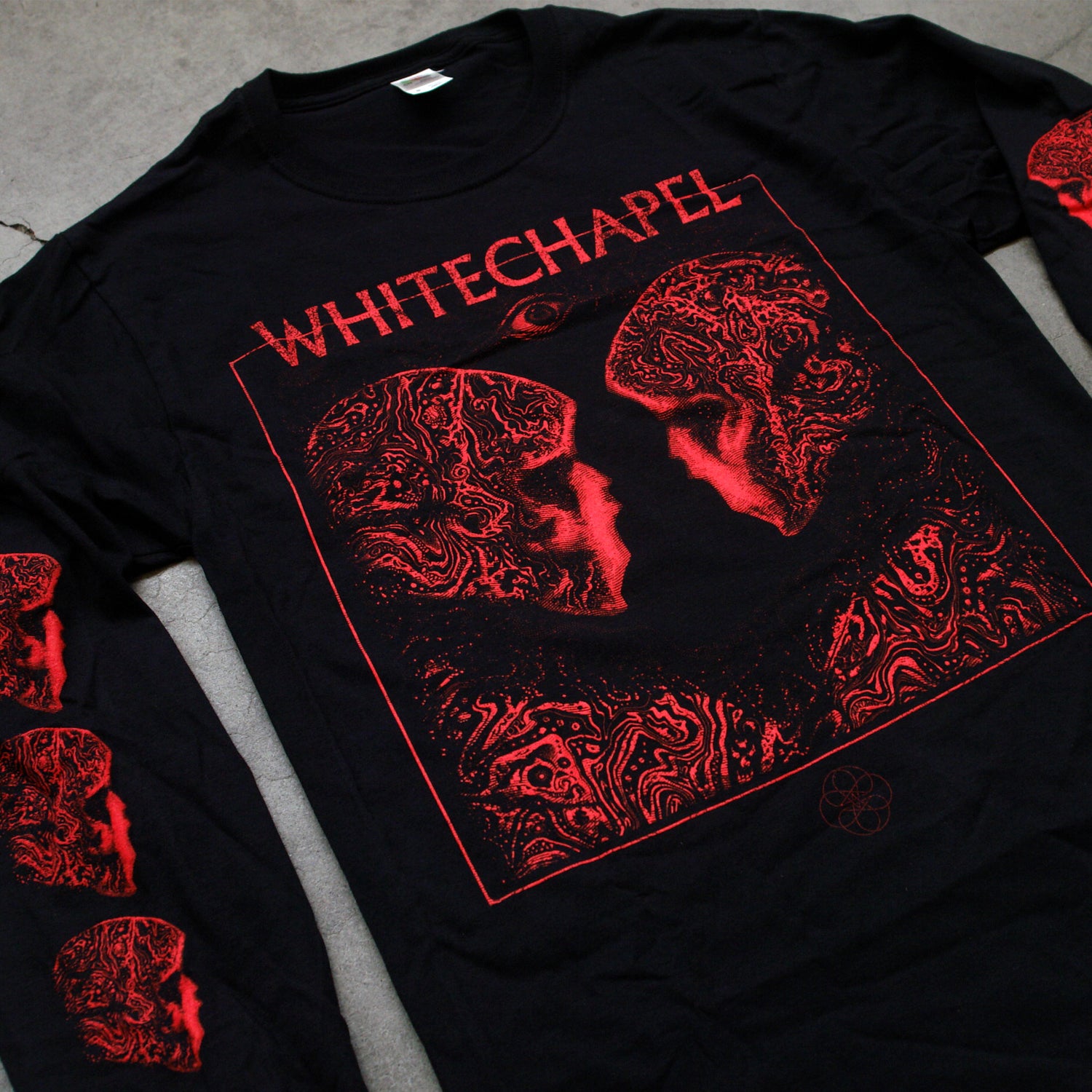 close up Image of the front of a black longsleeve on a grey concrete background. The front shows two skulls/skeletal figures from the shoulders up looking at one another- made up of red and black oil. This is outlined in a thin red square. Above the square in red text with the top line of the square going through the letters reads "whitechapel". Below the word "whitechapel" is a red image of an opened eyeball. Both sleeves have 3 of the skeletal figure heads on them, facing inwards.
