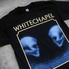 Load image into Gallery viewer, close up, angled image of the front of a black tee shirt on a concrete background. The front shows two skulls looking at one another- made up of blue and black static. The static is beneath them too. This is outlined in a tan square. Above the square in tan text reads &quot;whitechapel&quot;. 
