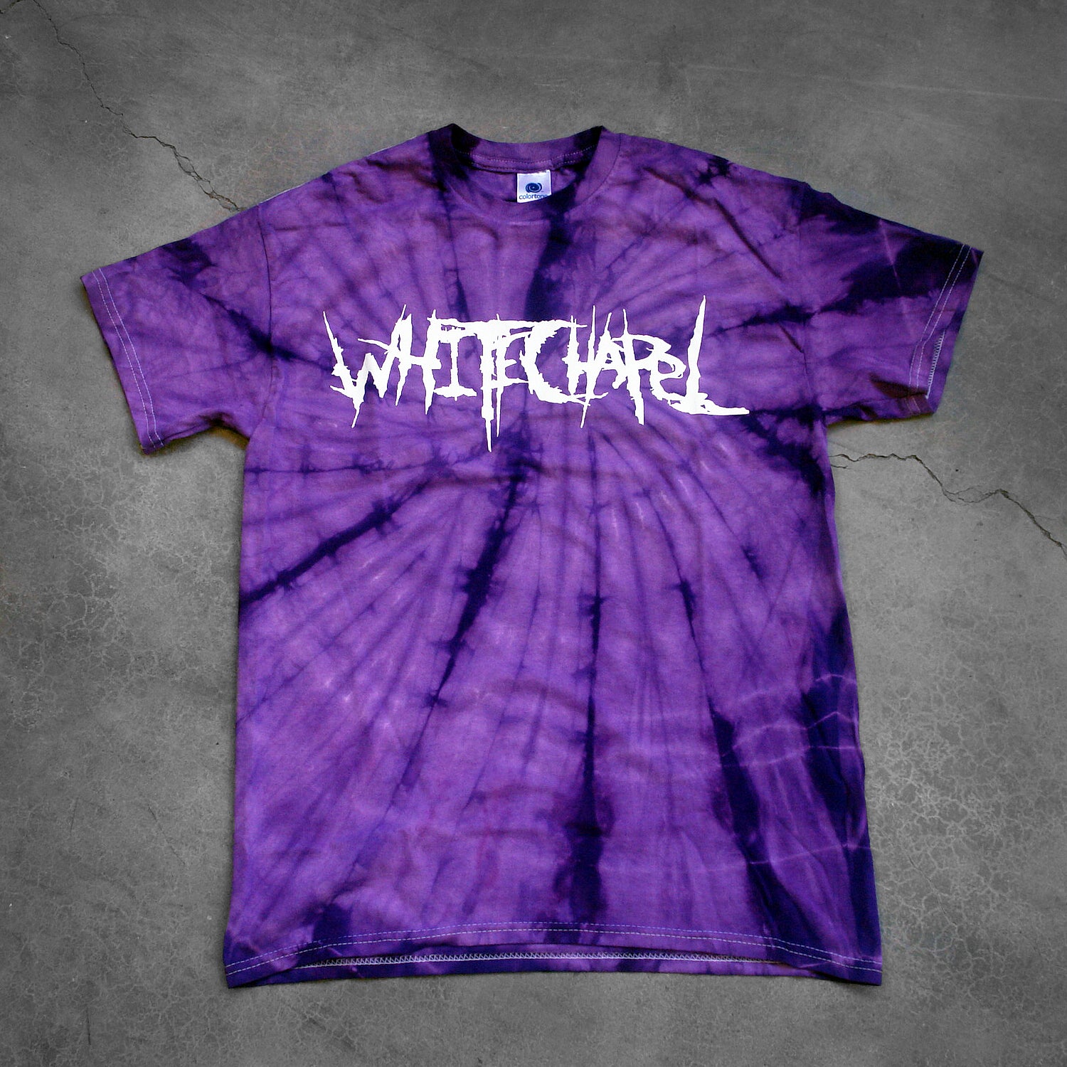 image of a purple spider tie dye tee shirt on a cracked concrete background. tee has front center chest print across in white that says Whitechapel
