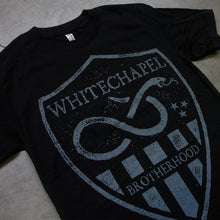 Load image into Gallery viewer, close up Image of a black tshirt against a grey concrete background. There is a graphic of a grey badge on the shirt, inside this badge in grey text reads &quot;whitechapel&quot;. Below this is a graphic of a snake with its tongue out. There are three small stars next to the snake. Below this are grey stripes, and in between the stripes in the middle reads &quot;brotherhood&quot;.
