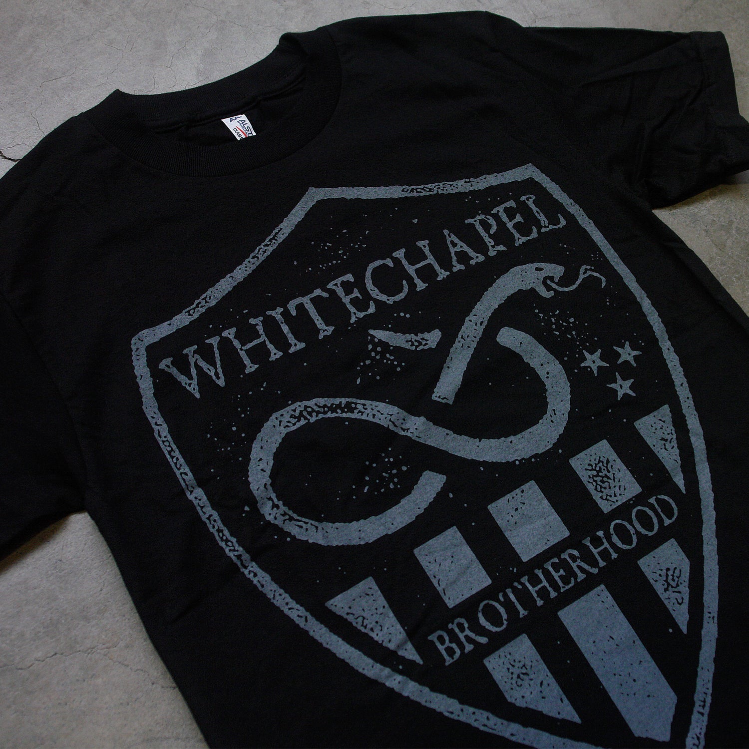 close up Image of a black tshirt against a grey concrete background. There is a graphic of a grey badge on the shirt, inside this badge in grey text reads "whitechapel". Below this is a graphic of a snake with its tongue out. There are three small stars next to the snake. Below this are grey stripes, and in between the stripes in the middle reads "brotherhood".
