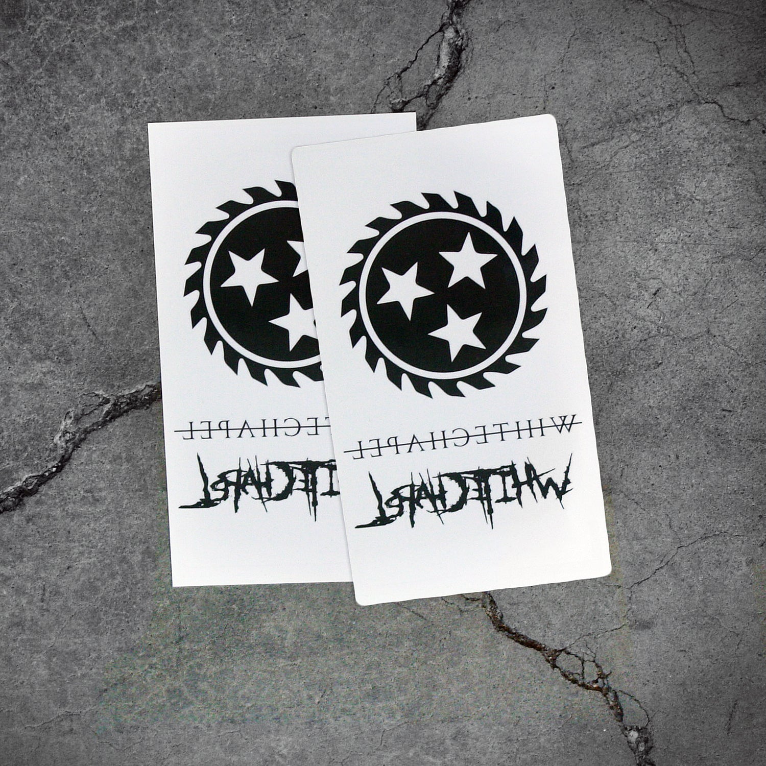 Image of two white pieces of paper with temporary tattoos on them against a grey concrete background. There is a black sawblade with three white stars in the center of it. There is the word whitechapel in black text with a thin black line through it, and the word whitechapel in black heavy metal style font. Both sheets are identical.