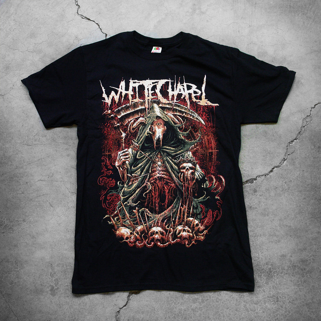 Image of the front of a black tshirt against a grey concrete background. the shirt features a red, white, black, and bluish gray graphic of a an animal skull with a third eye holding a scythe and wearing a cloak. Its ribs are showing. There are skull heads all around the monster. There is a red background behind the graphic.