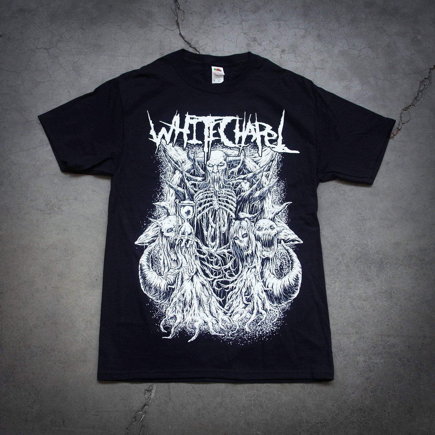 image of a black tee shirt laid flat on a cracked concrete floor. tee has full body print in white of a skeleton devil holding an hourglass and has several demons surronding it. at the top says whitechapel.