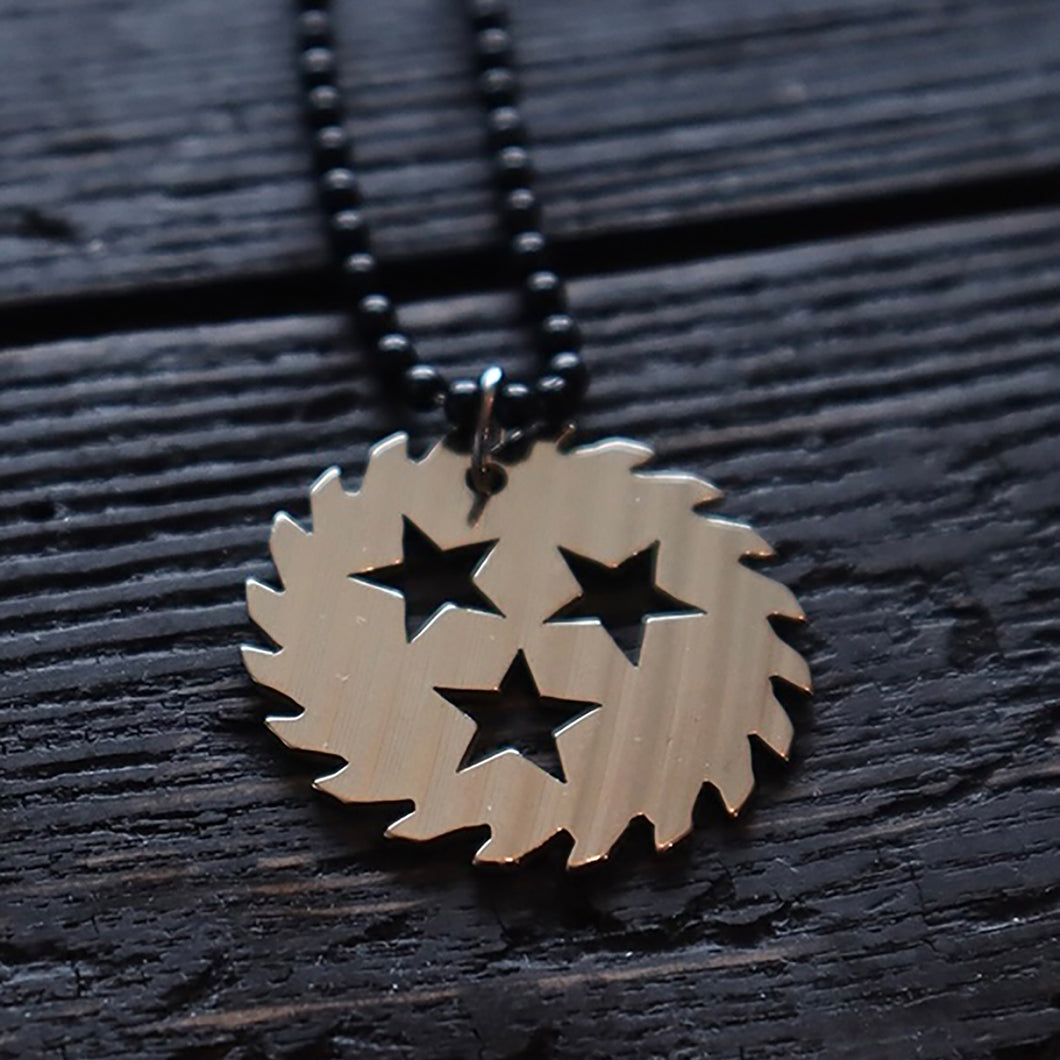 Image of a silver colored sawblade necklace against a black wooden background. The chain is made of black beads and has a silver metal sawblade with three stars cut out in the center of it on it. 
