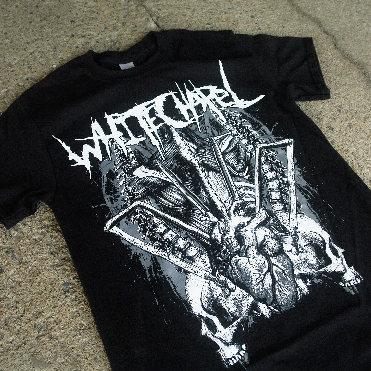 Image of the front of a black tshirt against a grey concrete background. The front of the shirt says "whitechapel" across the chest in white heavy metal style font. Below that is a white, grey, and black graphic of spines, a heart, bones, and two skeleton skulls, one facing towards the left, and the other mirrored, facing towards the right. 