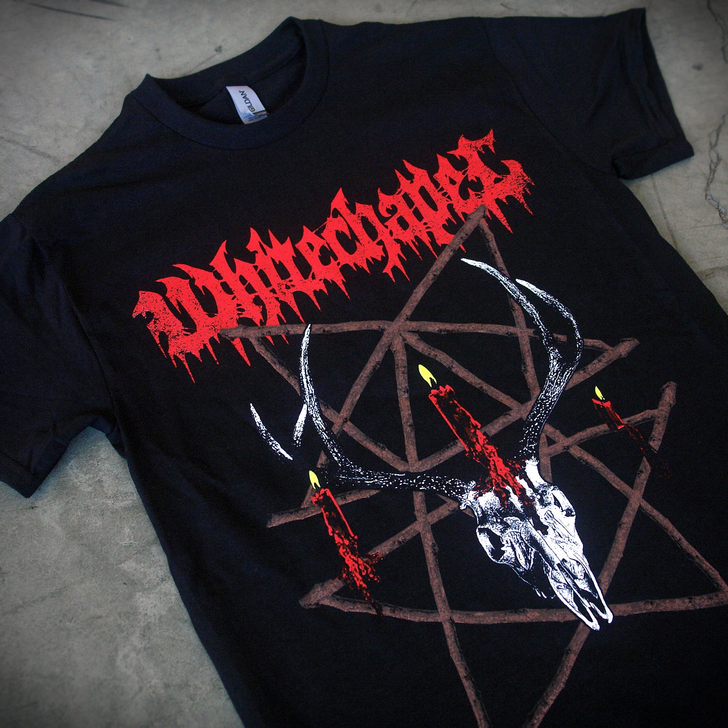 close up, angled image of a black tee shirt laid flat on a concrete floor. front of tee has full body print. at the top in red says whitechapel. below is a deer skull with a burning candle and a pentagram made out of sticks.